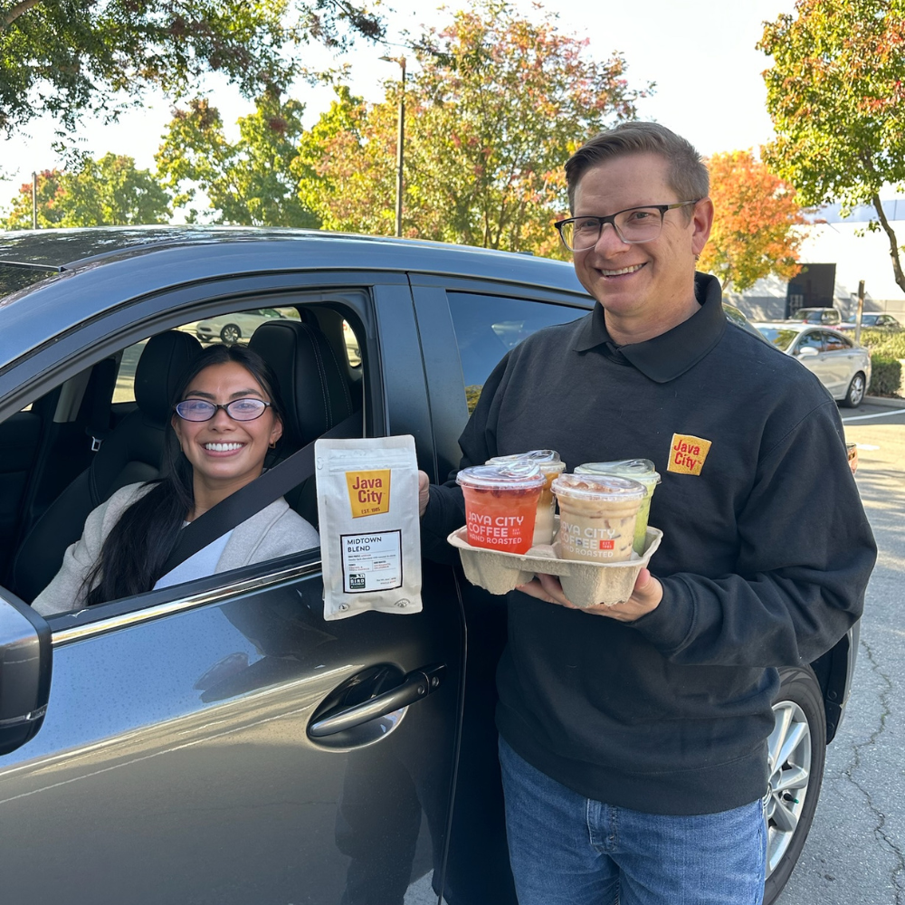 Man holding food containers and a bag, standing next to a car with a woman inside, in an outdoor setting.