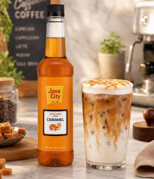 A bottle of Java City Caramel Syrup stands next to a glass of iced caramel latte on a marble countertop in a coffee shop setting.