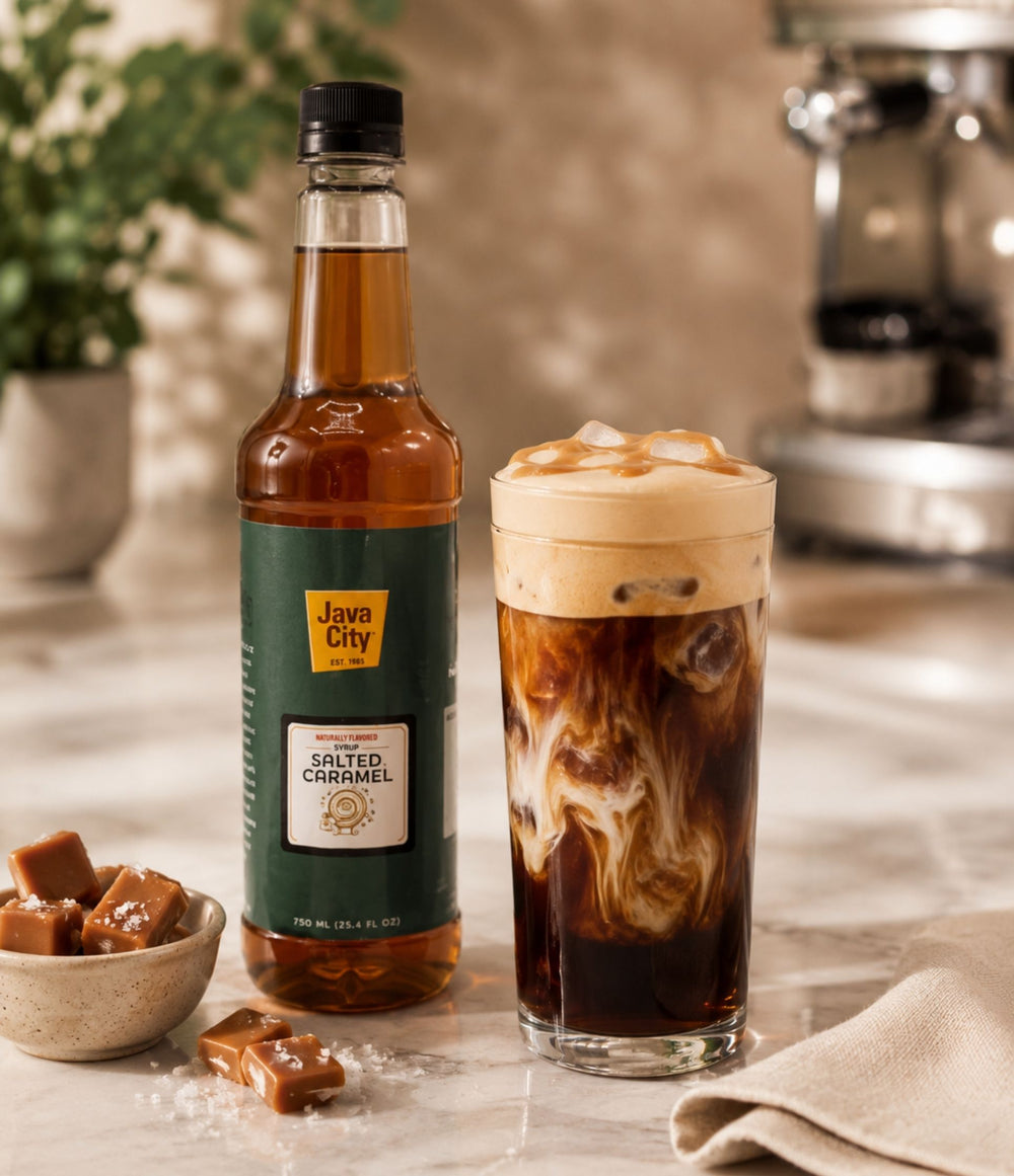 A bottle of Java City Salted Caramel syrup stands next to a tall glass of iced coffee with caramel swirl, set on a countertop with caramel pieces and a coffee machine in the background.